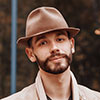 A guy with a stubble beard, brown fedora, and light work jacket stands outside on site, facing the camera; background is blurred. - Barndominium Kansas