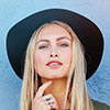 Individual with long blonde hair, black wide-brim hat, and olive green shirt smiles at camera; light blue background behind. - Barndominium Kansas